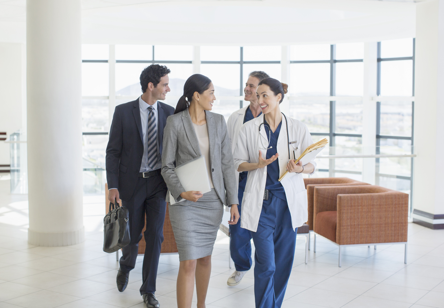 A group of four professionals, two wearing business attire, and two wearing medical attire, walk down a hall talking